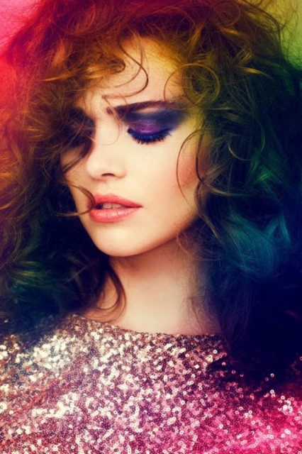 Close-up portrait of a woman with curly hair and dramatic purple eye makeup, wearing a sequined top with a colorful light effect.