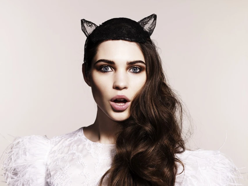 Woman poses in a studio with a dark cat-ear headband, white lace top with feathered shoulders, and dark wavy hair against a light background.