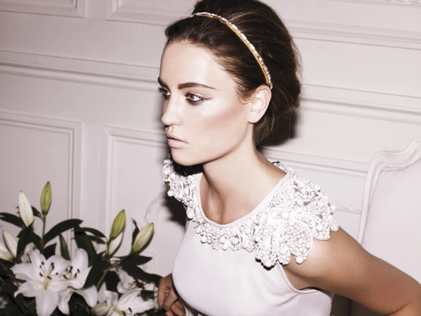 Brunette woman poses in a white dress with an ornate collar, accentuated with lily flowers and soft lighting against a paneled wall.
