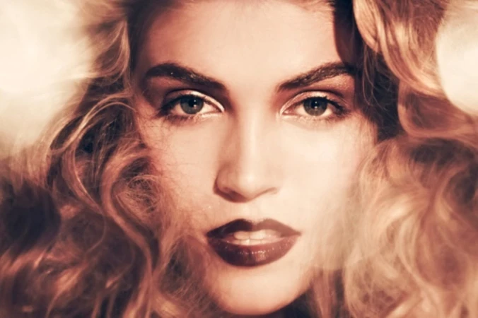Close-up portrait of a woman with voluminous blonde hair and dark lips, featuring make-up by Andriani Vasiliou.