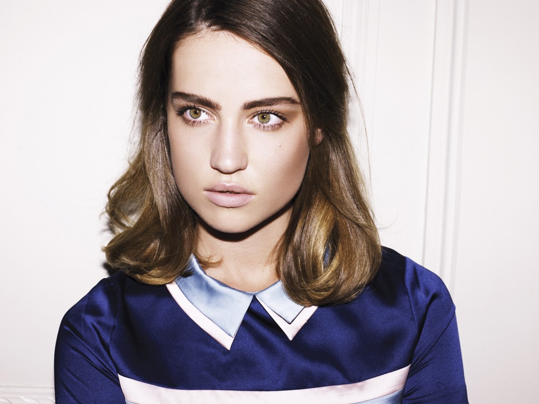 Close-up of a young adult woman with brown hair and eyes, wearing a navy and light blue collared dress, looking off-camera against a white wall.