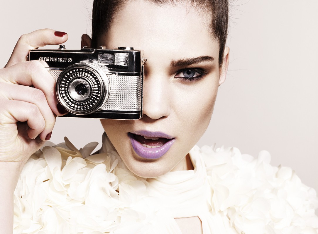 Woman in floral top holds a vintage Olympus Trip 35 camera up to her face, showcasing a close-up portrait indoors.