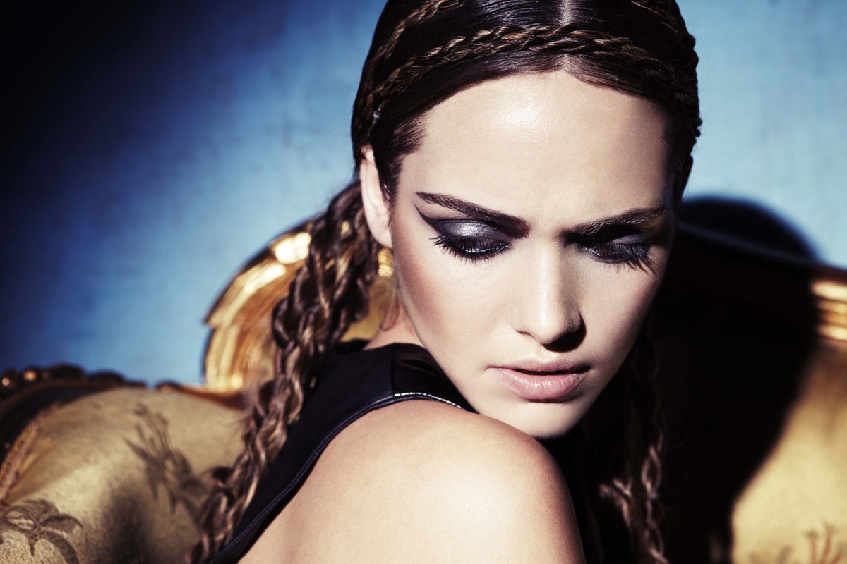 Close-up portrait of an adult female model with braided hair, dramatic eye makeup, and dark clothing, seated in an ornate armchair.