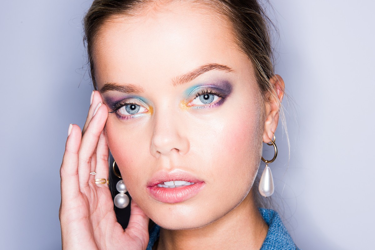 Close-up portrait of a fair-skinned woman with dramatic eye makeup, gold hoop earrings and pearl drops in a studio setting.