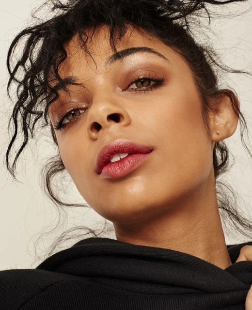 Close-up portrait of an adult female with dark curly hair wearing a black hooded sweatshirt, captured in soft lighting against a neutral background.