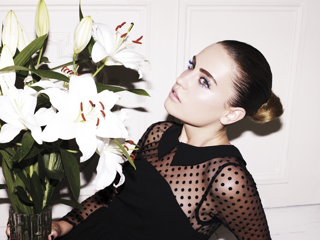 Portrait of an adult woman with fair skin and brown hair styled in a bun, wearing a black polka-dot dress, and posing next to white lilies.