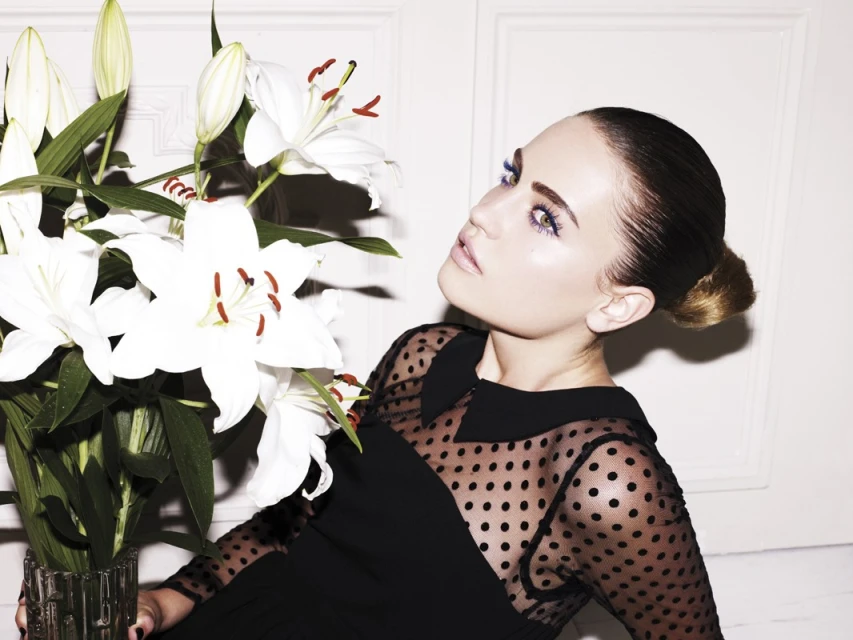 Portrait of an adult woman with fair skin and brown hair styled in a bun, wearing a black polka-dot dress, and posing next to white lilies.