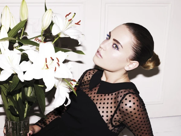 Portrait of an adult woman with fair skin and brown hair styled in a bun, wearing a black polka-dot dress, and posing next to white lilies.