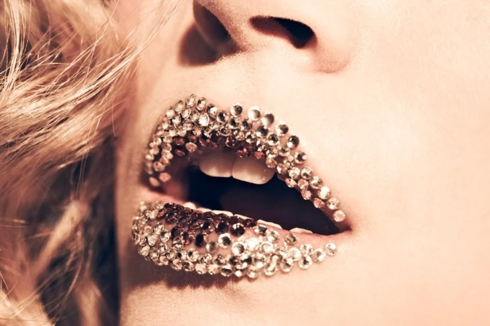 Close-up studio shot of a fair-skinned adult female with blonde hair, teeth visible, and glittering jewels adhered to her lips.