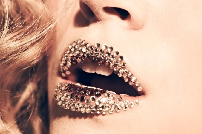 Close-up studio shot of a fair-skinned adult female with blonde hair, teeth visible, and glittering jewels adhered to her lips.