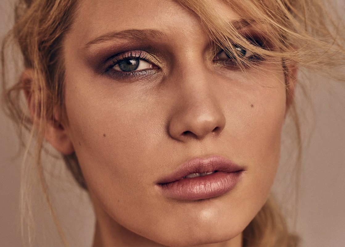 Close-up beauty portrait of a woman with smoky eye makeup and blonde hair framing her face against a neutral background.