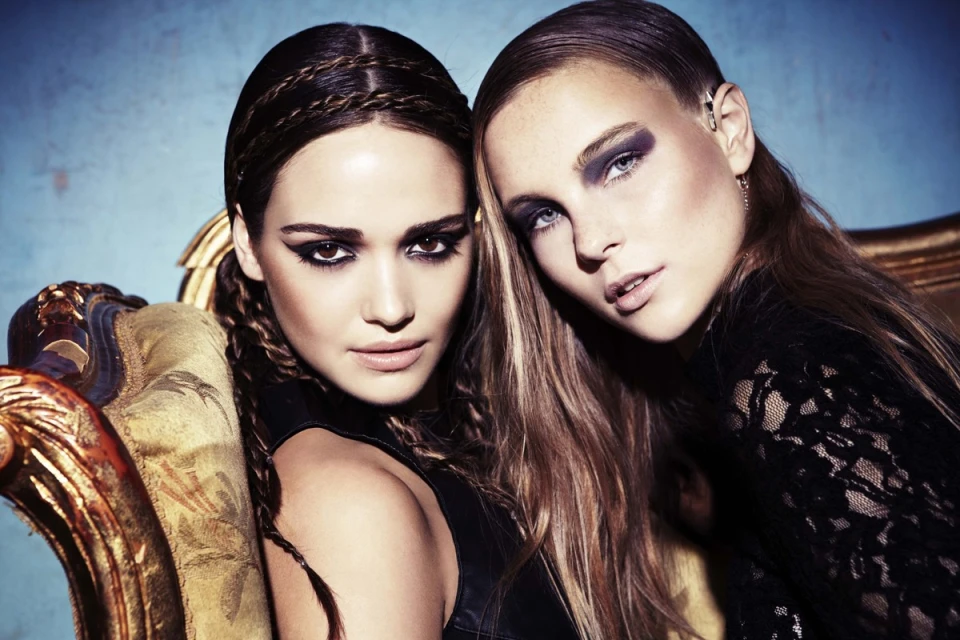 Two fashion models pose closely together, showcasing dark eye makeup and braided hairstyles, one resting on a golden ornate chair.