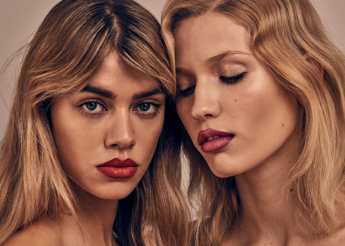 Close-up portrait of two young adult women with blonde hair and red lipstick posing indoors in a beauty and fashion context.