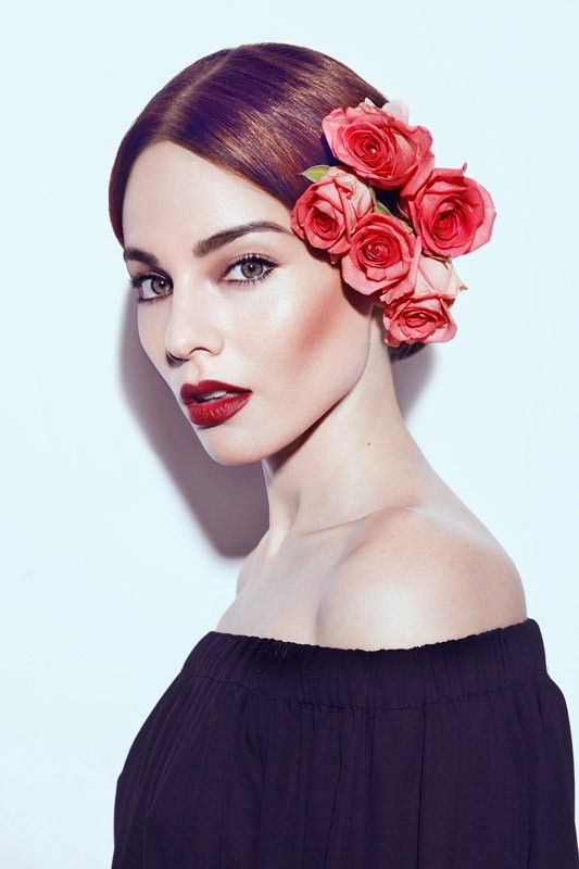 Woman wearing dark off-shoulder top with red roses in her hair against a bright white background