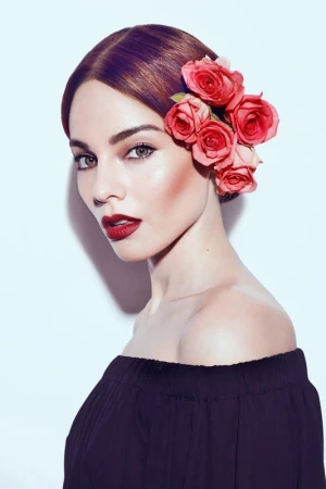 Woman wearing dark off-shoulder top with red roses in her hair against a bright white background