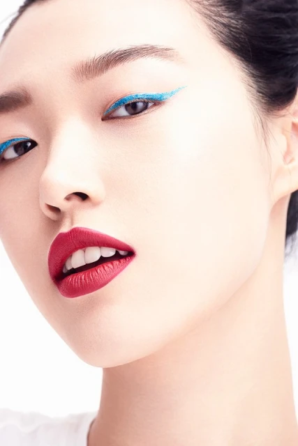 Close-up of Asian woman with red lipstick and blue eyeliner against a white background in a beauty style portrait.