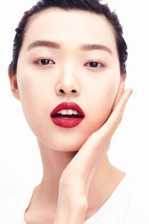 Close-up portrait of a woman with bold red lipstick touching her cheek against a clean white background