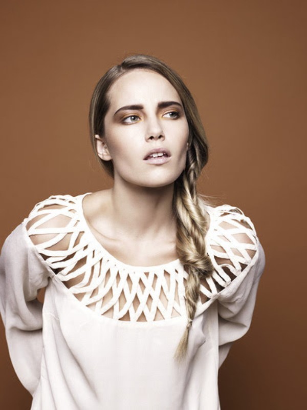 Portrait of a woman with a side braid wearing a white cutout top against a solid brown background, styled by Andriani Vasiliou.