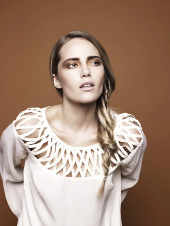 Portrait of a woman with a side braid wearing a white cutout top against a solid brown background, styled by Andriani Vasiliou.