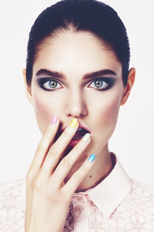 Close-up portrait of a woman with dramatic eye makeup and multicolored nail polish against a clean white backdrop in a photo studio.