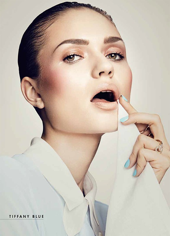 Close-up studio shot of a model with open mouth touching her lower lip with light blue painted nail and wearing a blue shirt.