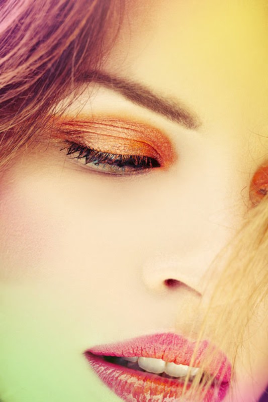Close-up of a woman's face with vivid eye makeup and lips. Her blonde hair partially covers her face. Soft, filtered light is present.