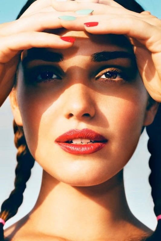 Close-up portrait of a woman with red lipstick, braided hair, and multicolored painted fingernails blocking the sunlight.