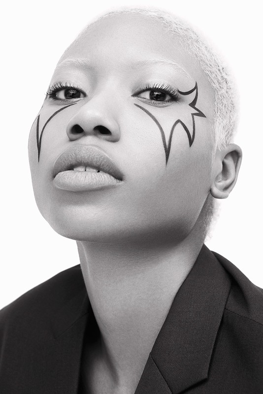 Close-up black-and-white portrait of a woman with a jacket and makeup designs around her eyes, set against a plain white backdrop.
