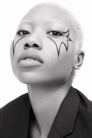 Close-up monochrome portrait of a person with short blonde hair and graphic eyeliner art makeup wearing a dark jacket.