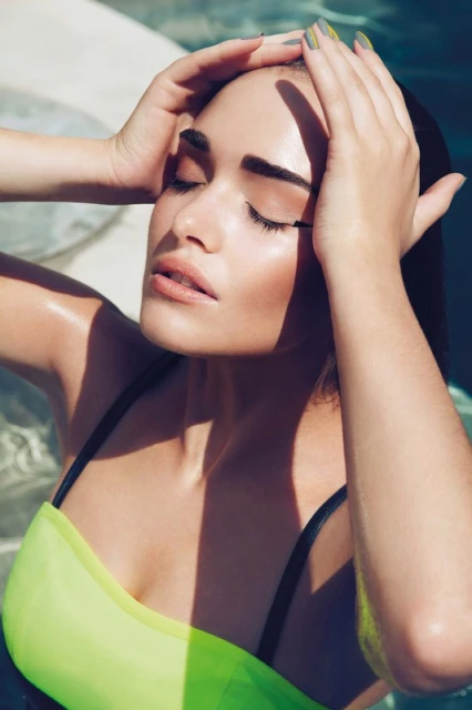 Close-up of a woman in a bathing suit with her eyes closed and her hands on her head, near water on a sunny day with casting shadows.
