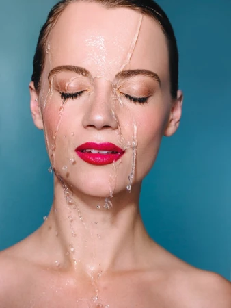 Close-up of a woman's face with eyes closed and water running down her face, set against a blue background for a fresh beauty concept.