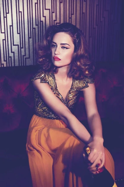 Glamorous adult woman in golden top and orange pants poses indoors holding a champagne bottle against a geometric background.