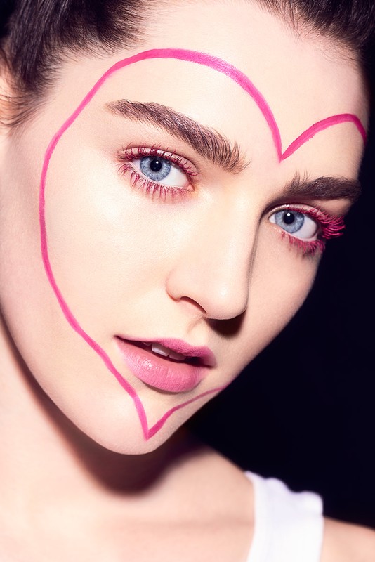 Close-up portrait of a young woman with pink heart makeup around her face and eyelashes against a dark backdrop, beauty shot.