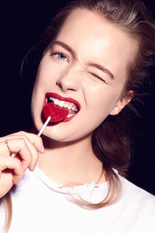 Close-up of a young adult woman winking, biting a heart-shaped lollipop, wearing a white t-shirt against a dark background.