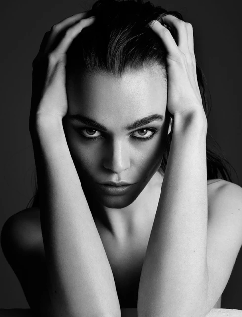 Black and white portrait of an adult woman with dark hair and makeup, looking directly at the camera with her hands near her head.
