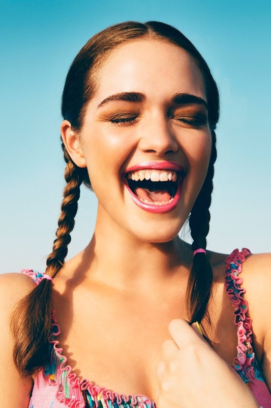Close-up of smiling young woman with pigtails against a clear blue sky, wearing a pink floral top, eyes closed, mouth open in laughter.