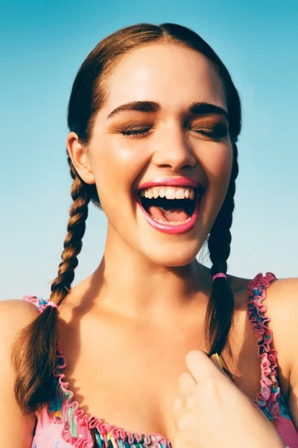 Close-up of smiling young woman with pigtails against a clear blue sky, wearing a pink floral top, eyes closed, mouth open in laughter.