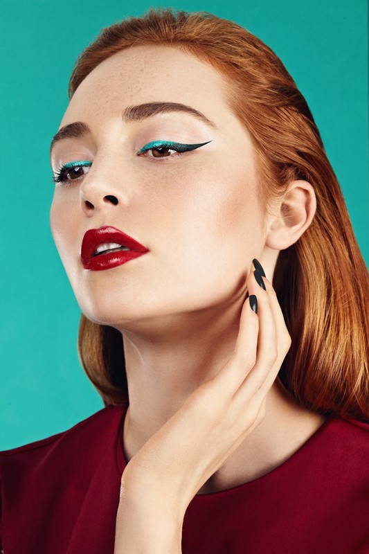 Portrait of a red-haired woman with red lipstick and blue eyeliner, posing with a hand near her face against a blue background.