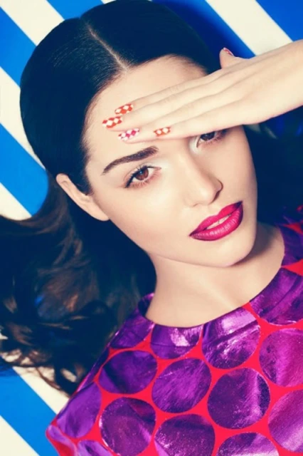 Close-up of a woman with dark hair wearing a polka-dot top, shielding her eyes with a hand, set against a blue and white striped backdrop.