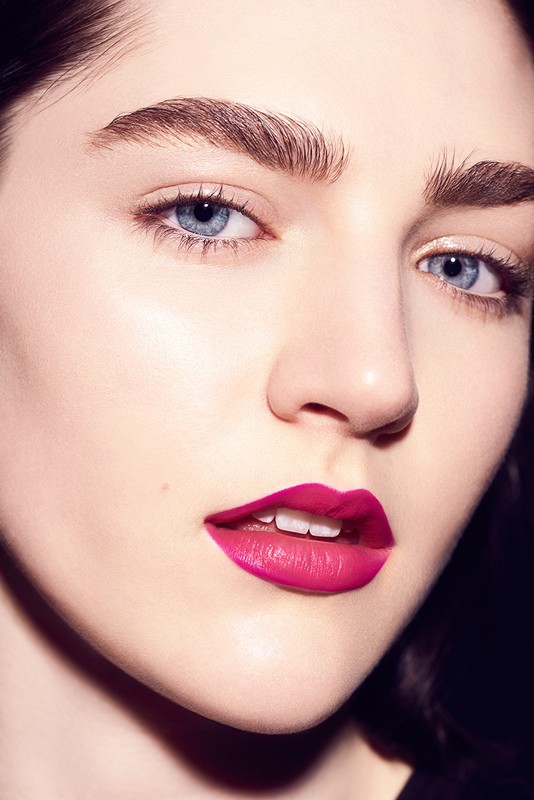 Close-up portrait of a young woman with blue eyes and magenta lipstick against a dark background, showcasing beauty and makeup.