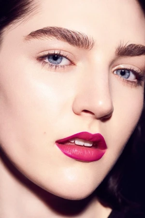 Close-up portrait of a young woman with blue eyes and magenta lipstick against a dark background, showcasing beauty and makeup.