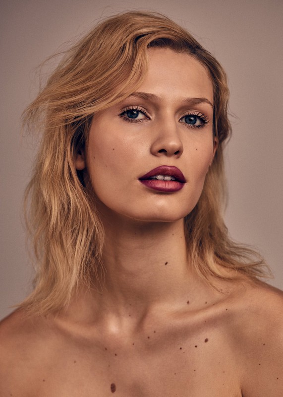 Close-up portrait of a blonde woman with dark lipstick and freckles against a neutral background.