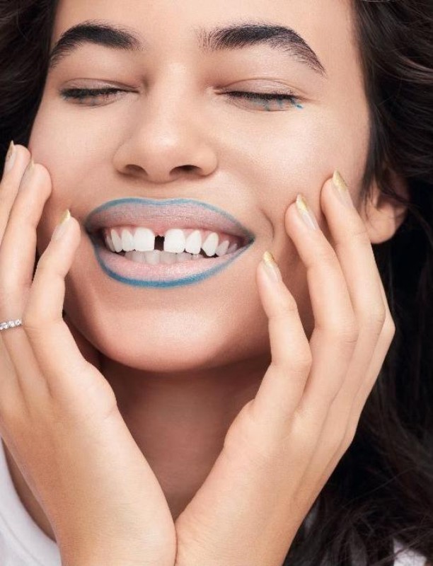 Close-up of a woman with closed eyes smiling with teal lipstick and a gap between her teeth, with her hands touching her face.