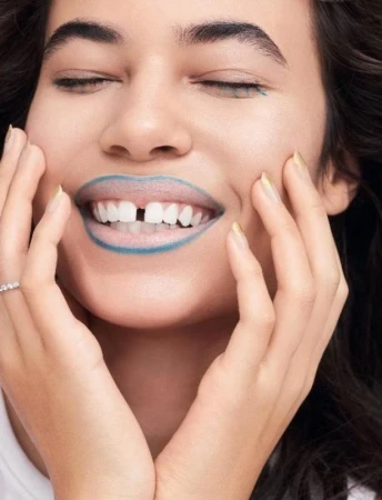 Close-up of a woman with closed eyes smiling with teal lipstick and a gap between her teeth, with her hands touching her face.