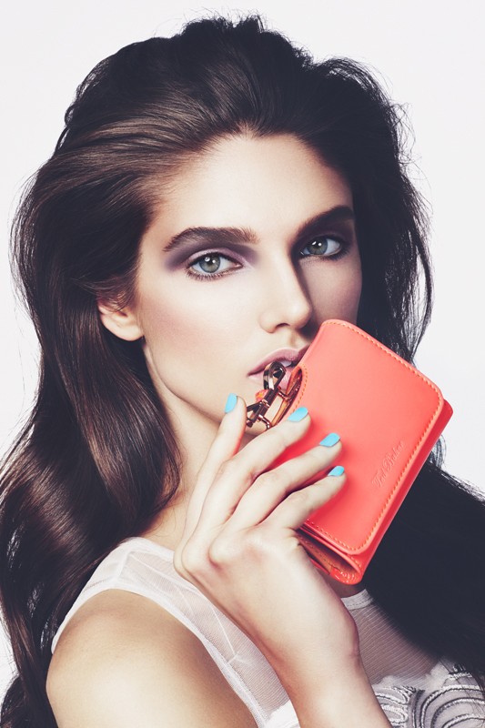 Woman in white top poses in photo studio holding an orange wallet to her mouth with hand showing turquoise nail polish.
