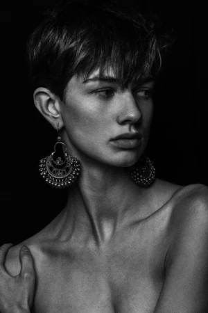 Monochrome portrait of a person with short textured hair wearing ornate metallic earrings against a dark background