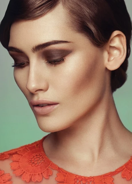 Close-up portrait of an adult female with brown eye makeup, styled dark hair, and an orange lace top against a gradient background.