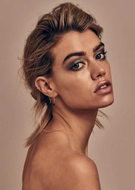 Close-up beauty shot of a blond woman with green eye shadow posing in front of a neutral backdrop inside a photo studio.
