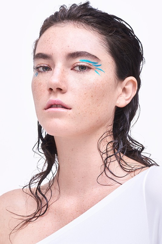 Close-up of a woman with wet dark hair and blue graphic makeup, posing against a stark white background in a beauty shot.