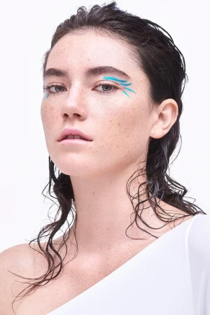 Close-up of a woman with wet dark hair and blue graphic makeup, posing against a stark white background in a beauty shot.